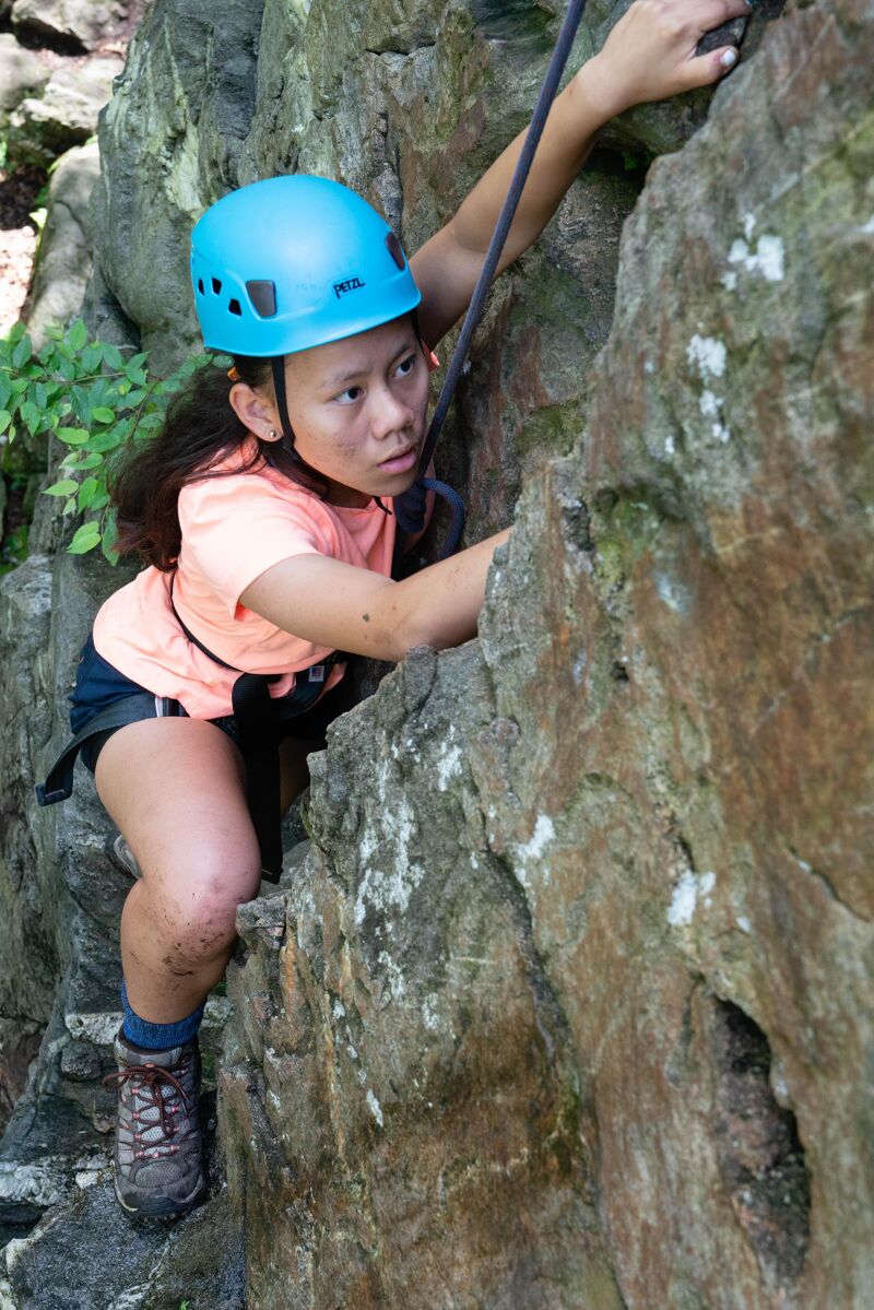 A young woman is rock climbing outdoors. She is wearing a blue helmet, an orange t-shirt, and dark shorts. She is gripping the rock face with her hands and feet, and there is a rope attached to her harness. The rock is grey and textured, and there are some green plants visible in the background. She seems focused and determined.