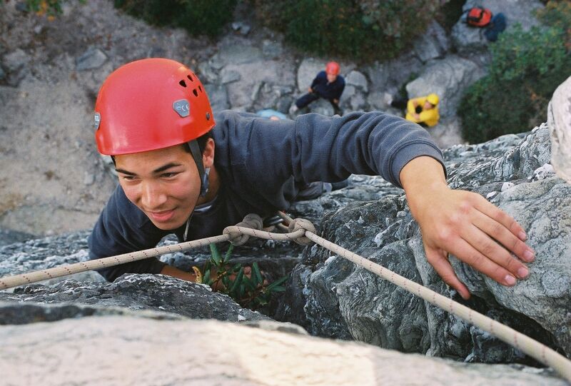 A man in a red helmet is rock climbing, focused and determined. He's gripping the rock face with one hand and a rope is visible, suggesting he's secured for safety. The setting appears to be outdoors, with other people and climbing gear visible in the background, indicating a group activity or a climbing area.