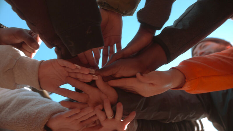 The image shows a group of people with their hands stacked on top of each other, forming a circle. The hands are of various skin tones, suggesting a diverse group. The composition is from a low angle, looking up at the hands against a blue sky. The focus is on the hands, symbolizing unity, teamwork, and collaboration. The overall impression is positive and conveys a sense of togetherness and shared purpose.