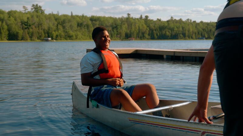 A young man wearing a white t-shirt and an orange life vest sits in a canoe on a lake. He is smiling and appears relaxed. The canoe is in the water near a wooden dock. The background features a lush green forest and a partly cloudy sky. Another person is partially visible on the right side of the image.